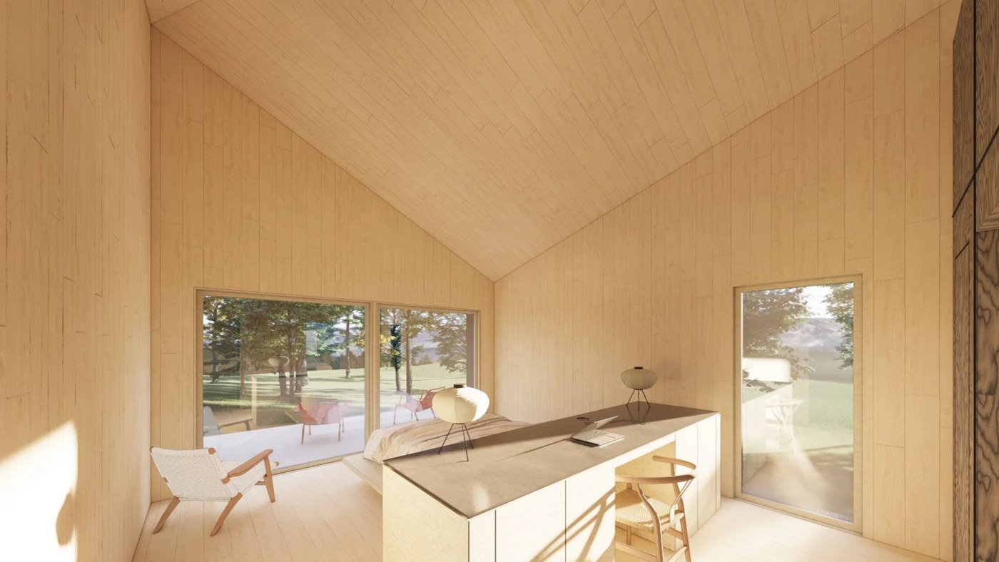 The island desk under the pitched ceiling, wide windows opening to the deck and meadow